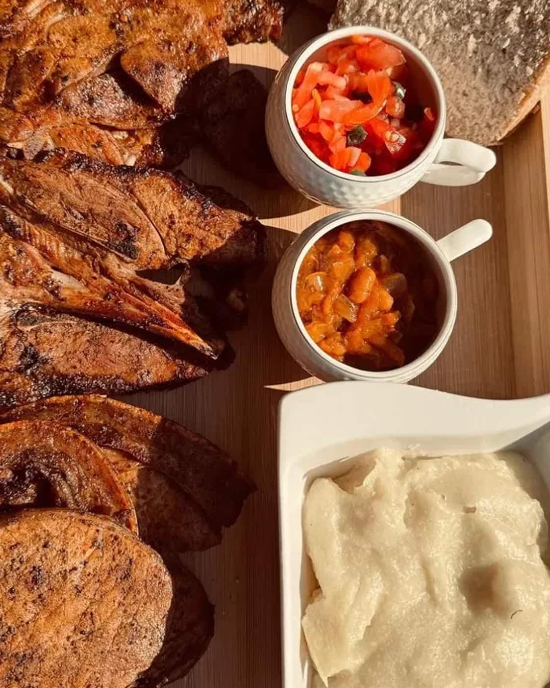 Grilled pork chops and ribs on a wooden board with a dish of phuthu, tomato salsa, chakalaka and thick-sliced bread