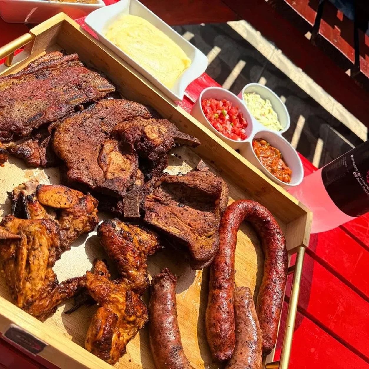 A coil of boerewors, beef ribs, chops and grilled chicken wings laid out on a wooden board, with pap and salads alongside