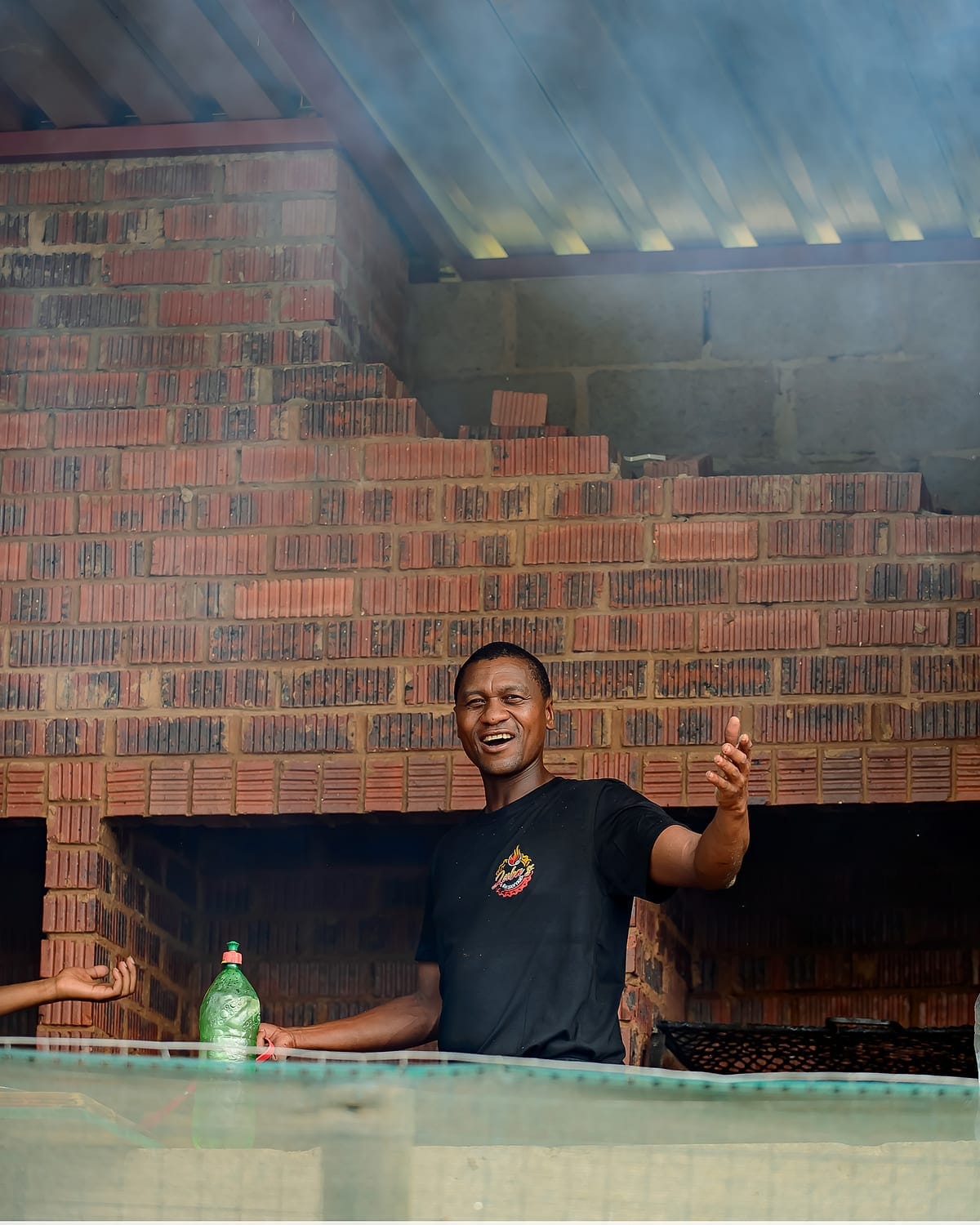 A man in a Zaba's Shisanyama shirt laughing behind the brick braai as smoke rises off the coals